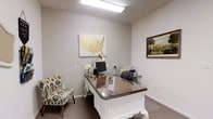 This single-story space presents a thoughtfully designed private office that balances professional functionality with decorative charm. The room is finished with neutral gray walls and carpeted flooring, creating a calm and focused atmosphere suited for professional consultations or administrative work.

The centerpiece of the office is an ornate white desk with carved detailing and a smooth, glossy surface, lending an elegant contrast to the otherwise understated décor. A dark executive chair sits behind the desk, accompanied by a monitor, printer, and a gold-toned adjustable desk lamp. A patterned accent chair in bold blue and yellow tones is positioned near the desk, offering a welcoming spot for visitors.

Wall décor includes a large framed map of the United States rendered in warm gold tones, serving as a notable focal point, alongside a framed landscape photograph on the adjacent wall. Overhead fluorescent lighting provides consistent illumination throughout the space. The textured ceiling adds subtle architectural interest. Overall, the office conveys a professional yet personable environment, well-suited for client-facing or administrative use.