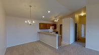 This single-story home showcases an open-concept layout that seamlessly connects the kitchen and living areas, creating a spacious and functional environment. The vaulted ceiling rises prominently throughout the shared space, enhancing the sense of openness and airflow. Recessed lighting and a ceiling fan provide practical illumination and comfort, while an elegant chandelier adds a decorative focal point above the dining or living area.

The kitchen is defined by warm-toned, rich wood cabinetry that provides ample storage, complemented by a white range and built-in range hood. A peninsula breakfast bar extends into the open living space, offering additional counter space and a casual seating area. The kitchen features tile flooring, which transitions naturally into the plush, neutral-toned carpet of the adjoining living area.

The living space is generous in scale, with clean white walls that lend a bright, neutral backdrop adaptable to various décor styles. A hallway visible beyond the kitchen suggests additional rooms further into the home. Overall, the space presents a practical yet welcoming atmosphere, well-suited for everyday living and entertaining.