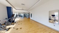 Located on Atlantic Avenue in Virginia Beach, this fitness facility offers a generously sized, single-floor layout designed to support a comprehensive workout experience. The space is defined by its clean, modern aesthetic, with light hardwood flooring throughout and crisp white walls that create a bright, energizing atmosphere.

The expansive exercise room is the centerpiece of the facility, housing a wide array of strength and cardio equipment, including treadmills, ellipticals, rowing machines, cable machines, free weights, and adjustable benches. Large mirrors line sections of the walls, enhancing the sense of openness, while multiple flat-screen televisions provide entertainment throughout workouts. A standout design feature is the striking artistic ceiling mural — a fluid, nautical-inspired motif rendered in deep blue and silver tones — that adds a distinctive coastal character to the space. Recessed and cove lighting further elevate the ambiance.

Adjacent to the main gym floor is a dedicated studio room, offering an open, uncluttered space ideal for yoga, stretching, or group classes. This room benefits from large windows that invite abundant natural light and views of the surrounding Virginia Beach streetscape. Built-in shelving provides organized storage for yoga mats, stability balls, and other accessories. Together, the two areas form a well-rounded, thoughtfully designed fitness environment.