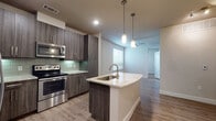 This single-floor residence showcases a thoughtfully designed open-concept layout with contemporary finishes throughout. The kitchen serves as the centerpiece, featuring dark wood-tone cabinetry, light granite countertops, and a full suite of stainless steel appliances, including a French door refrigerator, electric range with microwave, and dishwasher. A prominent kitchen island with an undermount sink and brushed gold faucet doubles as a prep and gathering space, illuminated by stylish pendant lights overhead. A soft green glass tile backsplash adds a subtle accent to the kitchen's neutral palette.

Wood-look flooring extends throughout the main living areas, creating a cohesive and easy-to-maintain surface. The living area features a ceiling fan and receives natural light through windows fitted with blinds. A glass-paneled entry door with a sidelight further brightens the space. One area transitions to plush carpet, offering a softer feel underfoot.

The bathroom is well-appointed with a dark wood vanity, granite countertop, large mirror with overhead lighting, tile flooring, and a combination tub and shower. The overall design maintains a clean, modern aesthetic with neutral tones and quality finishes.