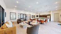 This single-floor space is designed with a contemporary aesthetic, combining an open-plan layout with a dedicated lounge area to create a versatile and inviting environment. The herringbone-patterned light wood flooring flows consistently throughout, grounding the space with a clean, modern foundation beneath crisp white walls and recessed lighting.

The open-plan kitchen, dining, and living area forms the heart of the space. The kitchen features dark espresso cabinetry, granite countertops, stainless steel appliances, and a large central island with bar seating, illuminated by globe pendant lights overhead. Adjacent to the kitchen, the dining zone accommodates multiple marble-topped round tables paired with richly upholstered burnt-orange chairs, adding warmth and color. Floor-to-ceiling French doors line one wall, flooding the area with natural light and providing direct access to an outdoor terrace.

The main living area offers an eclectic mix of seating, including a leather sofa, boucle chairs, and a stone-topped coffee table, anchored by a woven area rug. Black-framed glass partitions create a visual connection to adjoining spaces while maintaining definition. A separate, enclosed lounge provides an intimate setting with rounded upholstered chairs, a wall-mounted television, and a decorative wooden credenza, offering a quieter retreat within the overall layout.