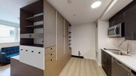 Located at 1500 Hancock St in Quincy, Massachusetts, this single-floor unit offers a modern, efficiently designed layout that maximizes functionality through intelligent use of space. The interior is anchored by a striking freestanding built-in storage unit crafted in warm wood tones, featuring a combination of open shelving, sliding cabinet doors, and drawers with matte black hardware. This central piece acts as a room divider, separating the living area from the rest of the unit while providing generous storage on multiple sides. The living area is finished with light-toned wood-look flooring and bright white walls, creating a clean and airy ambiance. Natural light enters through a window, adding warmth to the space. The kitchenette is outfitted with dark cabinetry, white quartz-style countertops, a textured tile backsplash, and stainless steel appliances including a microwave, induction cooktop, sink with a pull-down faucet, and a dishwasher — all thoughtfully compact yet fully functional. The bathroom features classic white subway tile in the walk-in shower, a mosaic herringbone tile floor, chrome fixtures, and accessibility-friendly grab bars, reflecting a practical and clean design aesthetic throughout.