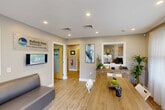 Located at 598 Bullocks Point Ave in East Providence, Rhode Island, this single-floor leasing office serves as the welcoming hub for Bullocks Point Village, an apartment community managed by The Simon Companies. The interior is thoughtfully designed with warm wood-look flooring, neutral gray walls, and recessed lighting throughout, creating a bright and professional atmosphere.

The main reception area doubles as a meeting space, featuring a large wood conference table surrounded by modern chairs, a comfortable seating area, and a reception desk tucked into a windowed alcove. Sheer white curtains filter abundant natural light from large windows, and contemporary abstract artwork adds a decorative touch to the walls.

Two private offices provide dedicated workspace for leasing staff. One office is furnished with a rich wood executive desk, an ergonomic chair, and bold abstract wall art, while a circular mirror and natural greenery add character to the space. A connecting hallway links the offices and includes access to additional utility areas.

The exterior of the property is equally inviting, with vibrant seasonal landscaping, mature trees, and well-maintained grounds surrounding the brick residential buildings of the community. The office entrance features a clear view of the Bullocks Point Village signage, establishing a strong sense of arrival for prospective and current residents.
