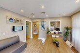 Located at 598 Bullocks Point Ave in East Providence, Rhode Island, this single-floor leasing office serves as the welcoming hub for Bullocks Point Village, an apartment community managed by The Simon Companies. The interior is thoughtfully designed with warm wood-look flooring, neutral gray walls, and recessed lighting throughout, creating a bright and professional atmosphere.

The main reception area doubles as a meeting space, featuring a large wood conference table surrounded by modern chairs, a comfortable seating area, and a reception desk tucked into a windowed alcove. Sheer white curtains filter abundant natural light from large windows, and contemporary abstract artwork adds a decorative touch to the walls.

Two private offices provide dedicated workspace for leasing staff. One office is furnished with a rich wood executive desk, an ergonomic chair, and bold abstract wall art, while a circular mirror and natural greenery add character to the space. A connecting hallway links the offices and includes access to additional utility areas.

The exterior of the property is equally inviting, with vibrant seasonal landscaping, mature trees, and well-maintained grounds surrounding the brick residential buildings of the community. The office entrance features a clear view of the Bullocks Point Village signage, establishing a strong sense of arrival for prospective and current residents.