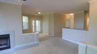 This single-floor space showcases an open-concept layout that seamlessly connects the living and kitchen areas. The main living space is finished with plush wall-to-wall carpeting in a neutral beige tone, complementing the warm tan wall color and crisp white crown molding that frames the room with a clean, classic detail. A set of French doors along one wall allows natural light to flow into the space while providing a direct view of the greenery outside, enhancing the overall brightness and openness of the interior. A side window further supplements the natural light. Track lighting is mounted near the window wall, offering additional illumination when needed. The kitchen area is partially visible and separated from the living space by a raised white half-wall counter, creating a natural visual division while maintaining the open feel of the floor plan. White cabinetry is visible in the kitchen, contributing to a clean and cohesive aesthetic. White baseboards throughout the space provide a sharp contrast to the carpeting and wall color, adding a polished finishing touch. The overall ambiance is understated, light, and functional.