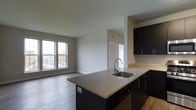 Unit 215 at 3610 Dillon St. is a single-floor residence that seamlessly integrates the kitchen and living areas into one cohesive open-plan layout. The kitchen is appointed with dark espresso cabinetry that provides a striking contrast against the light stone-finish countertops. A peninsula with an undermount sink serves as both a functional workspace and a natural dividing point between the kitchen and the broader living space, offering the potential for casual seating or additional counter use. Dark lower appliances, including a dishwasher, are integrated cleanly into the cabinetry design. The adjacent living area is bright and airy, benefiting from a large double-pane window that draws in generous natural light and offers exterior views. Smooth, light-toned hardwood-style flooring extends throughout the open area, contributing to a clean and modern aesthetic. The neutral gray walls and white ceilings provide a versatile backdrop suitable for a variety of interior styles. A doorway visible from the main living area suggests access to additional private spaces within the unit. Overall, the space delivers a modern, functional atmosphere with thoughtful design details.