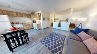 This single-story unit in Santa Fe, New Mexico, offers a well-planned layout that maximizes every square foot across its combined living spaces. The open-concept kitchen and living area form the heart of the unit, unified by durable wood-look luxury vinyl plank flooring that runs throughout. The kitchen is outfitted with warm, rustic wood cabinetry, a white refrigerator, dishwasher, and a light-toned countertop, complemented by a compact dining area positioned beside a large vertical-slat window. A ceiling fan overhead and track lighting provide both comfort and functionality. The living area flows naturally from the kitchen, providing room for seating and everyday use. A textured popcorn ceiling is consistent throughout the unit, adding a classic touch. A low half-wall partition subtly defines the transition between the living space and the generously sized primary bedroom. The bedroom comfortably accommodates a full-sized bed alongside additional furnishings, with neutral white walls, baseboard heating, and continued LVP flooring creating a calm, restful environment. Overall, the unit presents a clean, functional aesthetic well-suited for single-occupancy living in Santa Fe.