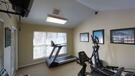 This single-floor fitness room offers a practical and functional workout space designed to accommodate essential cardio training. The room features neutral, light yellow walls that create a clean and unobtrusive atmosphere, complemented by carpeted flooring suitable for exercise equipment. A large window fitted with horizontal blinds allows natural light to fill the space, providing a bright and welcoming environment during daytime use. Overhead, a flush-mounted fluorescent light fixture ensures adequate illumination regardless of the time of day, while a ceiling-mounted security camera adds a layer of safety and oversight to the area. The room is equipped with a treadmill positioned near the window, an elliptical trainer, and a stationary bike, offering users a variety of cardio workout options. Two framed prints hang on the adjacent wall, adding a modest decorative touch to the otherwise utilitarian space. A ventilation grille in the ceiling supports air circulation, keeping the environment comfortable during workouts. Overall, the fitness room is straightforward and efficiently organized, providing residents or users with the key equipment needed for a productive cardiovascular exercise routine.
