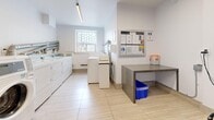Located at 740 Midland Ave in Toronto's Scarborough neighbourhood, this communal laundry facility is a clean, efficiently organized single-room space designed to serve building residents. The room features light wood-effect ceramic tile flooring and bright white walls, creating a fresh and hygienic atmosphere throughout.

A row of commercial-grade Huebsch front-loading washing machines lines one wall, accompanied by a matching set of dryers, providing ample laundry capacity. Instructional signage is prominently displayed above the machines to guide users. A top-loading washing machine and additional laundry units are positioned toward the rear of the room near a frosted window that allows natural light to filter in.

A sturdy metal folding table offers a dedicated surface for sorting and folding laundry, complemented by a blue recycling bin nearby. An information board mounted on the wall displays notices and building guidelines for residents. Two folding chairs and a payment kiosk are situated near the entrance, providing a convenient spot to wait and process payments.

Safety is well-addressed, with a fire extinguisher and electrical panel mounted near the entrance door. Linear LED lighting runs along the ceiling, ensuring consistent, bright illumination across the entire space. The room is well-ventilated and thoughtfully arranged to maximize usability and flow for everyday laundry needs.