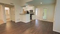This single-floor residence offers an open-concept layout that seamlessly connects the kitchen and living areas, creating a spacious and functional environment. The kitchen is outfitted with stainless steel appliances, including a refrigerator, microwave, and electric range, all complemented by white cabinetry and light-toned countertops. A peninsula with a breakfast bar extends into the living space, providing both additional prep space and casual seating. Rich, dark hardwood flooring flows throughout the main living area, adding warmth and visual continuity. Recessed lighting and under-cabinet fixtures illuminate the kitchen, while natural light enters through a glass-paned exterior door fitted with blinds, offering a view to the outdoors. The elevated ceiling above the kitchen area adds architectural interest and enhances the sense of openness. A carpeted room is visible through a doorway adjacent to the exterior door, suggesting a separate living or bedroom space. The overall ambiance is clean, modern, and move-in ready, with a neutral color palette that lends itself well to a variety of interior styles.