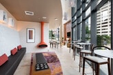 This contemporary lounge space is thoughtfully designed to balance comfort, style, and functionality. The main gathering area draws immediate attention with a striking orange suspended fireplace that serves as a bold focal point, complemented by a vibrant, multi-colored area rug and low-profile upholstered ottomans. Along one wall, a built-in dark leather banquette runs the full length of the space, backed by an expansive tufted white upholstered wall panel adorned with illuminated framed sculptural artwork. Pops of red accent pillows provide vivid contrast against the neutral tones. Floor-to-ceiling windows lined with teal-framed mullions flood the interior with natural light while offering views of the surrounding urban landscape. High-top tables and bar-height seating are positioned along the windowed perimeter, creating an inviting café-style zone. Overhead, warm gold-toned ceilings add a refined warmth throughout. A dedicated kitchenette area features warm cherry wood cabinetry, a light stone countertop, stainless steel appliances, an integrated sink, and a mosaic tile backsplash in earthy tones — ideal for serving and entertaining. Light travertine tile flooring ties the entire space together, creating a cohesive and polished aesthetic suitable for social gatherings and community events.