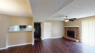 This single-floor residence offers a thoughtfully designed layout that balances comfort and functionality throughout. The living area serves as the heart of the home, anchored by a striking stone-surround fireplace set on a raised hearth, complemented by rich dark hardwood flooring and a ceiling fan overhead. A sliding glass door with vertical blinds floods the space with natural light and provides outdoor access. The open flow continues into the dining area, which features an elegant chandelier and connects seamlessly to the kitchen via a convenient pass-through counter. The kitchen is well-equipped with dark cabinetry, light granite-style countertops, a double stainless steel sink, and a matching suite of black appliances, including a full-size refrigerator. A set of double doors within the kitchen conceals additional storage or utility space. The bedrooms are carpeted for comfort, each outfitted with a ceiling fan and windows dressed with blinds that invite natural light and outdoor views. The bathroom features a dark vanity with a granite-style countertop, a large mirror with vanity lighting, and a separate toilet and bathing area. Textured ceilings and warm neutral wall tones carry throughout, giving the entire space a cohesive and welcoming ambiance.