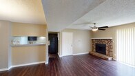 This single-floor residence offers a thoughtfully designed layout that balances comfort and functionality throughout. The living area serves as the heart of the home, anchored by a striking stone-surround fireplace set on a raised hearth, complemented by rich dark hardwood flooring and a ceiling fan overhead. A sliding glass door with vertical blinds floods the space with natural light and provides outdoor access. The open flow continues into the dining area, which features an elegant chandelier and connects seamlessly to the kitchen via a convenient pass-through counter. The kitchen is well-equipped with dark cabinetry, light granite-style countertops, a double stainless steel sink, and a matching suite of black appliances, including a full-size refrigerator. A set of double doors within the kitchen conceals additional storage or utility space. The bedrooms are carpeted for comfort, each outfitted with a ceiling fan and windows dressed with blinds that invite natural light and outdoor views. The bathroom features a dark vanity with a granite-style countertop, a large mirror with vanity lighting, and a separate toilet and bathing area. Textured ceilings and warm neutral wall tones carry throughout, giving the entire space a cohesive and welcoming ambiance.