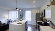 This single-floor space showcases a contemporary, open-concept kitchen and living area designed with a clean, minimalist aesthetic. The kitchen is a standout feature, centered around a sleek white island with a undermount sink, providing both functional workspace and casual gathering space. A linear pendant light hangs above the island, adding a modern focal point to the room.

The kitchen is well-equipped with a full suite of stainless steel appliances, including a refrigerator, range, microwave, and dishwasher, all complemented by crisp white cabinetry and light-toned countertops. Dark hardwood-style flooring runs throughout, creating a striking contrast against the white surfaces and light grey walls.

Natural light flows generously into the space through multiple windows fitted with white horizontal blinds, brightening the interior throughout the day. Recessed ceiling lighting supplements the natural light, ensuring the space remains well-lit at all hours. A glimpse of the adjoining dining area is visible, suggesting a seamless open floor plan that enhances the sense of spaciousness. The overall ambiance is clean, contemporary, and welcoming.