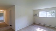 This single-floor space presents a straightforward and functional layout highlighted by neutral, light-colored walls that contribute to a bright and open atmosphere. The room visible in the image features wall-to-wall carpeting in a beige tone, providing a warm underfoot feel throughout. Natural light enters through a window on the far wall, which also houses a wall-mounted air conditioning unit, ensuring climate control during warmer months. A baseboard heating unit runs along the lower portion of the same wall, offering efficient warmth during cooler seasons. The walls are smooth and uniformly painted in a soft off-white tone, lending a clean and understated aesthetic. A partial wall or architectural divider is visible on the left side of the room, subtly defining the space without fully enclosing it. Electrical outlets and a wall switch are positioned conveniently along the walls. The overall ambiance is minimal and move-in ready, with practical climate control features built into the design. The space is well-suited for those seeking a functional, low-maintenance environment with essential amenities already in place.