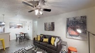 This single-story space is thoughtfully designed with an open-concept layout that connects the living, dining, and kitchen areas into one cohesive flow. The living room serves as the central gathering space, featuring dark hardwood-style flooring, a ceiling fan with integrated lighting, and textured ceilings. A bold black-and-white geometric area rug anchors the seating arrangement, complemented by urban-themed wall art that adds character to the bright, neutral walls with white paneling accents.

The kitchen is equipped with dark espresso cabinetry, stainless steel appliances including a refrigerator, range, and microwave, and a white countertop peninsula that doubles as a breakfast bar. Pendant lighting and track lighting illuminate the workspace effectively. Adjacent to the kitchen, the dining area accommodates a four-person table beneath a multi-arm chandelier.

Two bathrooms are present, each featuring dark-framed vanity mirrors, espresso cabinetry, and tub-shower combinations. Built-in shelving nooks near the bathrooms provide organized storage with basket systems, functioning as practical utility areas. Natural light enters through a window in the living area, contributing to the overall bright and welcoming ambiance throughout.