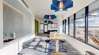 Located at 401 A St in Wilmington, Delaware, this single-floor communal living room is a thoughtfully designed lounge space that blends modern aesthetics with functional versatility. The room is characterized by crisp white walls, soaring ceilings, and an abundance of natural light streaming through large floor-to-ceiling windows that line the perimeter, offering open views of the surrounding cityscape and outdoor deck areas.

A series of striking blue geometric pendant light fixtures serve as the room's visual centerpiece, adding a bold, artistic accent to the bright, airy atmosphere. The flooring features a dynamic blue and grey patterned carpet that complements the cool, contemporary color palette found throughout.

The space is organized into distinct zones. A long communal high-top table with black stools provides a collaborative workspace or casual dining area, bathed in natural light from the adjacent windows. A lounge nook fitted with sleek blue sofas and a round coffee table creates a relaxed seating area ideal for conversation or unwinding. A vibrant lime-green upholstered bench built into a recessed nook near the entrance adds a playful pop of color.

A compact wet bar with a sink is neatly integrated along one wall, adding practical convenience. A glass door leads directly to an outdoor deck, seamlessly connecting the interior to the exterior environment. The overall ambiance is modern, welcoming, and energizing.