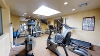 This fitness center offers a comprehensive workout environment designed to accommodate a range of exercise needs. The space is brightened by a large central skylight that floods the room with natural light, complemented by recessed ceiling lighting for consistent illumination throughout. Warm beige walls and carpeted flooring contribute to a comfortable, welcoming atmosphere.

The cardio area is well-stocked, featuring an upright stationary bike, a recumbent bike, and an elliptical trainer positioned for ease of use. A wall-mounted flat-screen television provides entertainment during workouts. Along the mirrored wall, a multi-station cable and strength machine offers versatile resistance training options, while a dumbbell rack with an organized selection of free weights supports additional strength exercises. The mirrored wall expands the perceived depth of the room and allows users to monitor their form during workouts.

A doorway visible at the far end suggests access to additional amenities or facilities. The overall layout is thoughtfully arranged to maximize usable floor space while maintaining clear pathways between equipment, creating an efficient and functional fitness environment.
