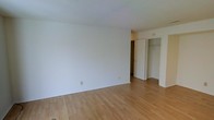 This single-floor space presents a straightforward and functional layout with a clean, minimalist aesthetic. The main living area is characterized by smooth white walls and light-toned hardwood flooring that runs throughout, lending warmth and continuity to the space. The neutral color palette creates a bright, open feel that serves as a versatile backdrop for a variety of interior styles.

A doorway visible from the main area leads to an adjoining space, suggesting a degree of separation between living and private areas. A closet with bi-fold or sliding doors is positioned along one wall, providing organized storage directly accessible from this area. The closet interior appears to include a hanging rod, offering practical storage solutions.

The ceiling is smooth and white, contributing to the overall sense of brightness. Electrical outlets are positioned at standard intervals along the baseboards, and the transition between rooms is clearly defined. The space is unfurnished, allowing prospective occupants to fully envision their own arrangement. Overall, the unit offers a tidy, neutral foundation well-suited for comfortable day-to-day living.