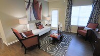 This single-floor space presents a thoughtfully designed interior that blends contemporary style with a warm, welcoming ambiance. The living area serves as the focal point of the space, showcasing rich, dark hardwood floors that anchor the room with a sense of warmth and sophistication. A large, bold abstract artwork in warm red and blue tones commands attention on the wall, adding a striking artistic element to the space. Natural light flows generously through the windows, which are dressed with elegant patterned curtains featuring a leafy motif, enhancing the room's visual depth. The living area is furnished with a clean-lined white sofa accented by decorative throw pillows in rust and black tones, complemented by a matching armchair in warm burnt orange. A sleek, dark coffee table sits atop a geometric patterned area rug in charcoal and white, adding texture and dimension to the floor. Candlestick holders on the coffee table and coordinating table lamps on either side of the sofa contribute to the cohesive, curated aesthetic. The overall design palette—combining neutral walls, dark wood accents, and rich warm tones—creates a stylish yet comfortable environment ideal for both relaxing and entertaining.