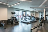Located at 3755 S Constitution Blvd in West Valley City, Utah, this fitness center offers a generously sized, single-floor exercise room designed to support a full range of workout routines. The space features a clean, modern aesthetic defined by crisp white walls, a dark speckled rubber flooring, and a striking combination of linear pendant and recessed lighting that illuminates the room with a bright, energizing atmosphere. Large windows along the exterior walls invite abundant natural light and provide outdoor views, enhancing the open feel of the space. The exercise room is comprehensively outfitted with a diverse selection of equipment, including multiple treadmills, a rowing machine, a stair climber, cable and pulley machines, a power rack with bench press, adjustable benches, and a full dumbbell rack. Floor-to-ceiling mirrors along one wall expand the sense of space and allow users to monitor their form during workouts. Structural columns are integrated thoughtfully throughout the layout without disrupting the flow of the space. A water fountain station with dual dispensers is conveniently positioned near the entry, and glass-paneled doors provide easy access. The overall design balances functionality with a polished, contemporary finish suited for serious fitness use.