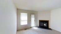 This multi-floor space presents a clean, straightforward design centered around comfort and functionality. The living area, visible in the provided images, is finished with neutral white walls and plush wall-to-wall carpeting that lends a soft, understated warmth to the room. A standout feature is the fireplace, framed by a warm wood surround and fitted with a classic black glass-door insert set against a dark slate hearth, serving as a natural focal point within the space.

Natural light enters generously through a large window dressed with horizontal slatted blinds, as well as through a glass-paneled exterior door — both positioned along the same wall, creating a bright and airy atmosphere. The door provides direct access to the outdoors, adding both convenience and connectivity to the exterior environment. Flush ceiling vents indicate a central HVAC system, contributing to year-round climate comfort.

The overall ambiance is calm and neutral, offering a blank canvas well-suited for a variety of interior styles. Spanning three floors in total, the property provides ample vertical space and room distribution beyond what is visible here.