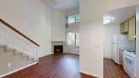 Located at 6210 Woodman Ave in Van Nuys, Los Angeles, this two-story residence offers a thoughtfully arranged layout spread across two floors. The main level features a bright, open living room distinguished by dramatically high ceilings that amplify the sense of space and airiness. Stacked windows flood the area with natural light and frame views of lush greenery outside, while rich hardwood-style flooring runs throughout. A tile-surround fireplace with a white mantel serves as a focal point, adding warmth and character to the living area. A carpeted staircase with a white-paneled railing and wood handrail leads to the upper level. The kitchen is open to the living area and features granite-style countertops, a breakfast bar pass-through, warm wood cabinetry, a gas range, and a double-basin sink beneath a window. Two full bathrooms are included in the home, each with a shower/tub combination enclosed by sliding glass doors and tile flooring. The upper floor provides additional living space with hardwood-style floors and access to a closet with built-in shelving. The overall ambiance is clean, bright, and functional, with a neutral palette throughout that offers a versatile canvas for personalization.