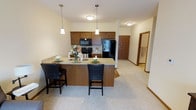 This single-floor residence offers a thoughtfully designed open-concept layout that seamlessly connects the kitchen and living areas, creating a bright and inviting atmosphere. The kitchen is a standout feature, showcasing warm medium-toned wood cabinetry that provides generous storage space. A granite-topped island with a breakfast bar serves as both a functional prep area and a casual dining spot, accommodated by two leather-upholstered bar stools. Pendant lighting hangs above the island, complementing the flush-mount ceiling fixtures to create a warm, layered lighting scheme. Black appliances, including a refrigerator, range, and over-the-range microwave, lend a sleek, modern contrast to the cabinetry. The living and dining areas feature soft, neutral carpet that adds warmth and comfort underfoot, while cream-toned walls keep the space feeling open and airy. White painted trim and baseboards provide clean, classic definition throughout. A hallway visible from the main living area leads to additional rooms, hinting at a well-organized floor plan. The overall ambiance is cozy yet polished, making this a comfortable and practical single-level living space.
