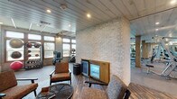 This well-appointed fitness facility combines rustic architectural character with a comprehensive range of modern exercise equipment. The space is defined by its distinctive light-colored stacked stone walls, exposed wood beams with timber support columns, and recessed lighting throughout, creating a warm yet energizing atmosphere.

The main workout floor is generously proportioned, accommodating a full row of cardio machines—including treadmills and ellipticals—along one windowed wall that allows ample natural light to fill the space. A dedicated strength-training area features cable machines, free weights, stability balls, and functional training equipment, while large mirrors line sections of the wall to support proper form.

Adjacent to the main gym floor is a comfortable lounge area finished with dark wood-plank-style flooring, upholstered chairs arranged around a woven coffee table, and a built-in beverage refrigerator. A water station is conveniently accessible nearby. Glass-paneled entry doors connect the lounge to the broader facility, reinforcing an open, cohesive layout.

Together, the industrial-rustic design elements, abundant equipment, and thoughtfully designed rest areas make this fitness center a functional and visually distinctive destination for exercise and recovery.
