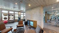 This well-appointed fitness facility combines rustic architectural character with a comprehensive range of modern exercise equipment. The space is defined by its distinctive light-colored stacked stone walls, exposed wood beams with timber support columns, and recessed lighting throughout, creating a warm yet energizing atmosphere.

The main workout floor is generously proportioned, accommodating a full row of cardio machines—including treadmills and ellipticals—along one windowed wall that allows ample natural light to fill the space. A dedicated strength-training area features cable machines, free weights, stability balls, and functional training equipment, while large mirrors line sections of the wall to support proper form.

Adjacent to the main gym floor is a comfortable lounge area finished with dark wood-plank-style flooring, upholstered chairs arranged around a woven coffee table, and a built-in beverage refrigerator. A water station is conveniently accessible nearby. Glass-paneled entry doors connect the lounge to the broader facility, reinforcing an open, cohesive layout.

Together, the industrial-rustic design elements, abundant equipment, and thoughtfully designed rest areas make this fitness center a functional and visually distinctive destination for exercise and recovery.