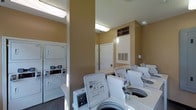 This communal laundry facility is thoughtfully designed to provide residents with a convenient and functional space for their laundry needs. The room features a row of top-load washing machines aligned along one side, complemented by stacked washer-dryer units that maximize the use of vertical space. The warm, neutral-toned walls create a welcoming atmosphere, while the bright fluorescent ceiling lighting ensures the space is well-lit throughout. HVAC vents integrated into the walls help maintain a comfortable environment even during heavy use. The facility is finished with a clean white aesthetic across all appliances, contributing to a tidy and organized appearance. A utility door provides additional access or storage, and an upper cabinet above the door offers extra storage space. The flooring is dark-toned, providing a practical contrast to the light-colored equipment. The layout is efficiently arranged to allow easy movement between machines, making the laundry process as smooth as possible. Overall, this single-floor laundry room delivers a practical, no-frills environment that prioritizes functionality and ease of use for its residents.