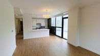 This single-floor space showcases a thoughtfully designed open-concept layout that seamlessly integrates the kitchen and living areas. The kitchen is outfitted with crisp white cabinetry, dark contrasting lower cabinets, and a clean white peninsula countertop that doubles as a casual dining or prep surface. Stainless steel appliances, including a microwave and range, are neatly integrated into the cabinetry, complemented by a pendant drum light that anchors the space with a warm, contemporary glow.

Warm-toned laminate hardwood flooring extends throughout, lending continuity and a sense of spaciousness across the entire open floor plan. The living area is generously proportioned, offering flexibility for various furniture arrangements.

A defining architectural feature is the floor-to-ceiling glass sliding door system framed in sleek black, which floods the interior with abundant natural light and provides direct access to an outdoor balcony. The cityscape views visible through the glass add a dramatic urban backdrop, enhancing the overall ambiance. The neutral white walls and clean lines throughout contribute to a bright, modern aesthetic suited to contemporary urban living.