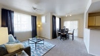 This single-floor space features an open-plan layout that seamlessly connects the living and dining areas, creating a sense of spaciousness and flow. The living area is anchored by plush wall-to-wall carpeting and centers around a glass-top coffee table set atop a geometric blue area rug, complemented by a ceiling fan that enhances comfort. Large windows dressed with vertical blinds and dark curtain panels line the walls, allowing an abundance of natural light to fill the space while offering privacy and light control. The dining area is positioned adjacent to the living space and is furnished with a dark wood dining table and matching chairs, accommodating several guests. Track lighting overhead provides focused illumination for the dining area. A sliding glass door fitted with vertical blinds leads to an outdoor area, bringing in additional natural light and offering a seamless indoor-outdoor connection. The warm, neutral-toned walls and light ceiling contribute to the welcoming, airy ambiance of the space. Overall, the layout is practical and well-lit, making it well-suited for both everyday living and casual entertaining.