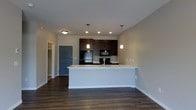 This single-floor space offers a thoughtfully designed open-concept layout that seamlessly connects the kitchen and living areas. The kitchen is a standout feature, showcasing rich dark wood cabinetry that extends to the upper wall, paired with black appliances including a refrigerator, range, and over-the-range microwave. A large peninsula with a light-toned countertop and an integrated sink serves as both a functional workspace and a natural dividing point between the kitchen and the main living area. Two modern pendant lights hang above the peninsula, adding focused illumination and a contemporary aesthetic touch. Recessed lighting in the kitchen ceiling further enhances the brightness of the space. The flooring throughout is a deep, dark hardwood that adds warmth and sophistication to the overall design. The walls are finished in a soft neutral tone, complementing the contrast between the dark cabinetry and light countertops. A teal-accented entry door is visible toward the rear, adding a pop of color and leading to what appears to be a closet or additional room. The layout is clean, open, and well-suited for everyday living, with a functional flow that makes the space feel welcoming and efficiently organized.