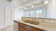 Located on Morand Alley in the heart of Cincinnati, OH, this urban residence blends industrial character with modern finishes. The open-concept layout is anchored by light hardwood flooring that runs throughout, while exposed structural beams and high ceilings lend a distinctive loft-style aesthetic. Large multi-pane windows bathe the living area in natural light and frame views of the surrounding downtown cityscape.

The kitchen is well-appointed with granite countertops, rich dark wood cabinetry, a subway tile backsplash, stainless steel appliances — including a refrigerator, electric range, microwave, and dishwasher — and a peninsula with pendant lighting that opens to the living space. A long hallway connects the main living areas to the private quarters, offering ample storage with three closets and a utility room.

The bedroom features a ceiling fan, exposed beam detailing, and a window that continues the urban views. Two full bathrooms serve the residence, one of which includes a soaking tub with granite-surround tile walls and polished chrome fixtures. The overall ambiance is bright and airy, successfully merging the raw texture of industrial architecture with clean, contemporary finishes throughout.