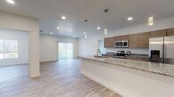 This well-designed single-floor unit offers a clean, contemporary aesthetic throughout. The open-concept layout connects the kitchen and living area seamlessly, creating a bright and airy atmosphere enhanced by recessed lighting and elegant pendant fixtures. The kitchen is a standout feature, equipped with stainless steel appliances including a range, microwave, and French door refrigerator, complemented by warm wood-toned cabinetry and granite countertops that extend across a generous island with a built-in sink. Light wood-look flooring runs consistently throughout the unit, adding warmth and visual continuity. The living area benefits from natural light via a sliding glass door fitted with vertical blinds, while the entryway provides a clean, welcoming transition into the space. The bedrooms are well-proportioned and bright, with large windows framing views of surrounding greenery and distant mountain ridgelines. The bathrooms are finished with granite-topped wood vanities, brushed gold fixtures, and clean white tile, with one bathroom offering a walk-in shower and another featuring a tub-shower combination. The overall ambiance is modern, neutral, and move-in ready, with consistent finishes that lend a cohesive and polished feel throughout.