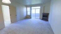 This two-floor space offers a comfortable and functional layout designed for everyday living. The main living area is a highlight of the space, featuring plush wall-to-wall carpeting that adds warmth underfoot. A built-in fireplace with a dark surround anchors one corner of the room, providing a cozy focal point. Natural light flows generously into the space through a combination of a standard window and a wide sliding glass door, which opens to an outdoor area and frames views of surrounding greenery. The sliding door also allows for easy indoor-outdoor access, enhancing the sense of openness. Smooth, neutral-toned walls and a flat ceiling with a ceiling fan contribute to the clean, understated aesthetic throughout. An arched architectural detail near the fireplace adds a subtle design touch. The room includes a wall-mounted heating unit beneath the window, ensuring year-round comfort. The overall ambiance is calm and move-in ready, with a neutral palette that provides a versatile backdrop for a variety of interior styles. The two-floor configuration offers additional living or sleeping space beyond the main level.