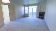 This two-floor space offers a comfortable and functional layout designed for everyday living. The main living area is a highlight of the space, featuring plush wall-to-wall carpeting that adds warmth underfoot. A built-in fireplace with a dark surround anchors one corner of the room, providing a cozy focal point. Natural light flows generously into the space through a combination of a standard window and a wide sliding glass door, which opens to an outdoor area and frames views of surrounding greenery. The sliding door also allows for easy indoor-outdoor access, enhancing the sense of openness. Smooth, neutral-toned walls and a flat ceiling with a ceiling fan contribute to the clean, understated aesthetic throughout. An arched architectural detail near the fireplace adds a subtle design touch. The room includes a wall-mounted heating unit beneath the window, ensuring year-round comfort. The overall ambiance is calm and move-in ready, with a neutral palette that provides a versatile backdrop for a variety of interior styles. The two-floor configuration offers additional living or sleeping space beyond the main level.