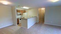 This single-floor residential space offers a functional and straightforward layout designed for comfortable everyday living. The heart of the home is an open-concept kitchen and living area, where a peninsula-style counter with a dark granite-look surface serves as a natural divider between the two spaces. The kitchen is equipped with white appliances, including a gas range and a full-sized refrigerator, complemented by warm honey-toned wood cabinetry that provides ample storage. An under-cabinet range hood and overhead fluorescent lighting keep the cooking area well-lit and practical. The adjacent living area features light neutral carpeting that extends throughout, creating a cohesive and cozy atmosphere. A hallway visible from the main living space leads further into the unit, hinting at additional rooms beyond. The entry area, partially visible at the end of the hall, includes a thermostat panel and a solid entry door. The overall palette of soft beige walls, light carpet, and warm wood tones gives the space a neutral, move-in-ready feel. The layout is efficient and well-organized, making it easy to navigate between the main living and kitchen zones.