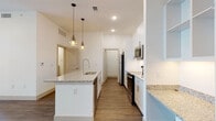 This single-floor residence offers a thoughtfully designed open-concept layout that seamlessly connects the kitchen and living areas. The kitchen serves as the centerpiece of the home, showcasing a large central island with light granite countertops, an integrated sink with a sleek pull-down faucet, and elegant pendant lighting with geometric cage-style fixtures. Crisp white cabinetry lines the walls, complemented by stainless steel appliances including a refrigerator, range, and microwave. A built-in desk nook with matching granite and open shelving provides a dedicated workspace. Warm hardwood-style flooring flows throughout the entire space, adding a cohesive and contemporary feel. The living area benefits from natural light streaming through large windows fitted with blinds, and a ceiling fan adds comfort overhead. A walk-in closet with built-in shelving offers generous storage. The bathroom is a refined retreat, featuring a frameless glass walk-in shower fully clad in large-format marble-look tile with veined detailing, brushed nickel fixtures, and matching marble tile flooring. Clean white walls and recessed lighting throughout create a bright, airy ambiance across the entire residence.