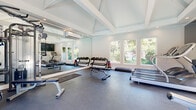 The Spring Creek Fitness Center at 7761 Greenback Ln in Citrus Heights, CA is a dedicated exercise facility designed to support a comprehensive workout experience. The space is housed within a single-room layout featuring a striking vaulted, coffered ceiling with exposed beams painted white, lending the room a sense of openness and height. Recessed lighting throughout ensures the space is well-lit at all times, while a wall-mounted mini-split HVAC unit provides climate control year-round.

A full wall of floor-to-ceiling mirrors dramatically expands the perceived depth of the room and is a key architectural feature, providing exercisers with proper form feedback during workouts. Large multi-pane windows along one wall invite abundant natural light and offer views of the surrounding greenery. French doors on an adjacent wall open directly to an outdoor pool area, further enhancing the connection to the outdoors.

The durable rubber tile flooring is practical and cushioned, ideal for high-impact fitness activities. Equipment includes multiple treadmills, elliptical trainers, a stationary bike, a multi-function cable machine, a weight bench, and organized dumbbell racks with a full range of free weights. A wall-mounted television and a water fountain add to the functionality of this well-appointed fitness space.