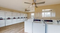 This single-story clothes care facility is designed to efficiently accommodate high-volume laundry needs. The space is outfitted with a generous selection of machines, including multiple top-load and front-load washers alongside an extensive row of stacked dryers lining the walls. Each machine features coin-operated controls, and laundry tip signage is posted for user guidance in both English and Spanish.

The interior is finished with durable wood-look flooring that runs throughout, complementing the neutral warm-beige wall tones and textured ceiling. Two ceiling fans are suspended overhead, promoting consistent air circulation throughout the facility. Natural light enters through several windows positioned along the walls, providing views of the surrounding exterior grounds and contributing to a bright, open atmosphere.

A folding table is available for convenience, along with a waste bin near the entrance. The layout is organized to allow comfortable movement between machines, with washing units centrally positioned and dryers arranged along the perimeter walls. The overall design prioritizes functionality and ease of use, making this facility a practical and well-maintained resource.