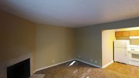 This single-floor space features a generously sized main room finished with wall-to-wall carpeting and a combination of warm tan and cool gray wall tones, creating a neutral, versatile interior palette. White baseboard trim lines the perimeter, providing a clean contrast against the painted walls. The standard-height ceiling is finished in white, contributing to a bright and open feel within the room. A doorway on the right wall opens to an adjacent area, suggesting a functional layout with connected living spaces. The transition from carpet to tile flooring is visible near the entryway, indicating a shift in room use or zone. Electrical outlets are positioned at standard intervals along the walls, offering practical utility throughout the space. The room's simple, unadorned design provides a blank canvas well-suited for a variety of furnishing and décor arrangements. Overall, the space presents a clean, move-in-ready interior with a straightforward layout and neutral finishes that offer broad appeal and flexibility for personalization.