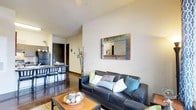 This single-floor space features an open-concept layout that seamlessly integrates the living and dining areas into one cohesive environment. The living area is anchored by a dark leather sofa dressed with a mix of patterned and solid accent pillows in teal, grey, and white tones. A rustic wooden coffee table sits atop a textured area rug, adding warmth to the space. A decorative mirror with a multicolored reclaimed-wood frame serves as a focal point on the main wall, complementing the neutral palette throughout. The ceilings are of a generous height, contributing to an open, airy feel. Polished concrete-style flooring runs throughout, providing a sleek, modern foundation. Adjacent to the living area, a dining space features a bar-height table paired with dark bistro-style stools, creating a casual yet functional eating area. A dark cabinetry unit nearby adds contrast to the light walls. Subtle decorative elements, including tall dried branches and small potted greenery, add organic texture to the interior. A ceiling fan overhead provides both function and a finishing touch to this well-appointed space.