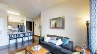This single-floor space features an open-concept layout that seamlessly integrates the living and dining areas into one cohesive environment. The living area is anchored by a dark leather sofa dressed with a mix of patterned and solid accent pillows in teal, grey, and white tones. A rustic wooden coffee table sits atop a textured area rug, adding warmth to the space. A decorative mirror with a multicolored reclaimed-wood frame serves as a focal point on the main wall, complementing the neutral palette throughout. The ceilings are of a generous height, contributing to an open, airy feel. Polished concrete-style flooring runs throughout, providing a sleek, modern foundation. Adjacent to the living area, a dining space features a bar-height table paired with dark bistro-style stools, creating a casual yet functional eating area. A dark cabinetry unit nearby adds contrast to the light walls. Subtle decorative elements, including tall dried branches and small potted greenery, add organic texture to the interior. A ceiling fan overhead provides both function and a finishing touch to this well-appointed space.