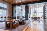This expansive, single-level lobby lounge is defined by a refined contemporary aesthetic that combines bold architectural elements with carefully curated design details. The space is anchored by soaring ceilings clad in warm wood slat paneling, which contrasts elegantly with deep navy and charcoal accent walls throughout. Large floor-to-ceiling windows dressed in sheer white curtains flood the interior with natural light while framing views of the surrounding urban skyline.

A dramatic focal point is established by a large-scale art installation featuring an arrangement of vinyl records mounted on a wall panel, adding a cultural and creative dimension to the space. Nearby, angular black structural columns create a bold geometric statement. The reception area features a striking striped marble countertop paired with sleek dark cabinetry and integrated LED lighting.

Multiple distinct seating zones are thoughtfully arranged throughout, including a lounge area centered around a built-in linear fireplace flanked by wood and dark-framed shelving, a game table with upholstered stools, high-top bar seating along the windows, and a casual sofa nook with side tables and greenery. Polished large-format stone flooring and patterned area rugs with gold detailing complete the sophisticated, cohesive atmosphere.