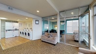This single-floor amenity space is thoughtfully designed to serve the practical and professional needs of its residents. The layout is open and efficiently organized, combining multiple functional areas in a cohesive, welcoming environment.

The laundry facility is prominently positioned and features a row of side-by-side washing machines and dryers, set against bright yellow-toned walls with informational postings, and finished with warm wood-look flooring for a clean, approachable feel.

Adjacent to the laundry area is a comfortable lounge space with carpet tile flooring in a neutral, textured pattern. A loveseat with an accent pillow provides a relaxed spot to wait, while the open layout keeps the area feeling airy and uncluttered.

A glass-enclosed business center is a standout feature of this floor, offering full transparency through floor-to-ceiling glass walls and sliding glass doors. Inside, workstations with chairs are arranged for focused productivity, and the room is accented with a bold teal wall that adds a vibrant pop of color. Natural light filters in through a window, keeping the workspace bright and inviting.

Recessed lighting throughout maintains consistent, even illumination across all areas. The overall design balances functionality with comfort, creating a practical yet pleasant communal environment.