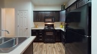 This single-floor residence offers a thoughtfully designed layout that balances comfort and functionality across its key living areas. The kitchen is a standout feature, showcasing rich dark espresso cabinetry that extends to the ceiling, light-toned countertops, and a full suite of black appliances including an electric range, over-the-range microwave, and refrigerator. Wood-look flooring ties the kitchen seamlessly into the adjacent dining area, which provides an intimate setting with a round marble-top table and access to colorful wall art and mirror accents that add depth to the space. The living area is welcoming and open, with light neutral walls, wood-look flooring, and a comfortable seating arrangement anchored by a dark leather sofa. The bedroom is carpeted for warmth and comfort, featuring a ceiling fan with light fixture, multiple windows dressed with blinds and sheer curtains that invite natural light, and ample space for furnishings. The bathroom is finished with stone-look tile flooring, a full tub-shower combination, and a built-in shelving unit for convenient storage. Throughout the residence, clean lines, neutral tones, and consistent design choices create a cohesive and inviting atmosphere.