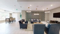 Located at 301 Continental Pkwy in Newport News, Virginia, this single-floor community space offers a well-appointed lounge and dining area designed with a contemporary aesthetic. The living room area is anchored by a striking decorative accent wall featuring a teal and wood-toned geometric pattern, serving as a sophisticated backdrop for a wall-mounted flat-screen television atop a sleek, gold-accented media console. Plush navy leather seating — including sofas and armchairs — is arranged around a sculptural coffee table and a textured area rug, while a pair of oversized circular pendant lights with brushed gold hardware provide warm, layered illumination throughout. The adjacent dining area benefits from an abundance of natural light streaming through large windows dressed in sheer white curtains, offering views of the surrounding landscape. A tall bar-height table surrounded by upholstered chairs serves as a functional gathering spot, complemented by vibrant large-format abstract artwork adorning the neutral-toned walls. Light, large-format tile flooring flows seamlessly throughout the space. Glass sliding doors connect to an additional amenity area beyond, enhancing the open, accessible feel of this inviting community hub.