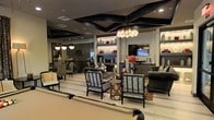 This sophisticated single-floor space makes an immediate impression with its striking interior design and carefully curated aesthetic. The open-plan layout connects a lounge area, a living zone, and a kitchen, creating a fluid and cohesive environment ideal for both entertaining and everyday use.

The ceiling is a standout architectural feature, adorned with bold, angular dark panels that add depth and drama to the space. Recessed lighting and a statement chandelier with layered glass shades provide warm, ambient illumination throughout.

The lounge area is furnished with patterned accent chairs in a geometric Greek key motif, complemented by a dark tufted sofa and small decorative side tables. A central display of tall decorative vases and floral accents adds visual interest and a refined touch.

Built-in shelving units with integrated lighting line the walls, showcasing curated décor pieces and glassware, flanking a wall-mounted flat-screen television that serves as a focal point for the living area.

The kitchen, visible in the background, features dark cabinetry, a decorative tile backsplash, and pendant lighting, maintaining the rich, moody color palette that defines the space. Overall, the interior exudes a contemporary, upscale ambiance with a cohesive dark-toned design language throughout.